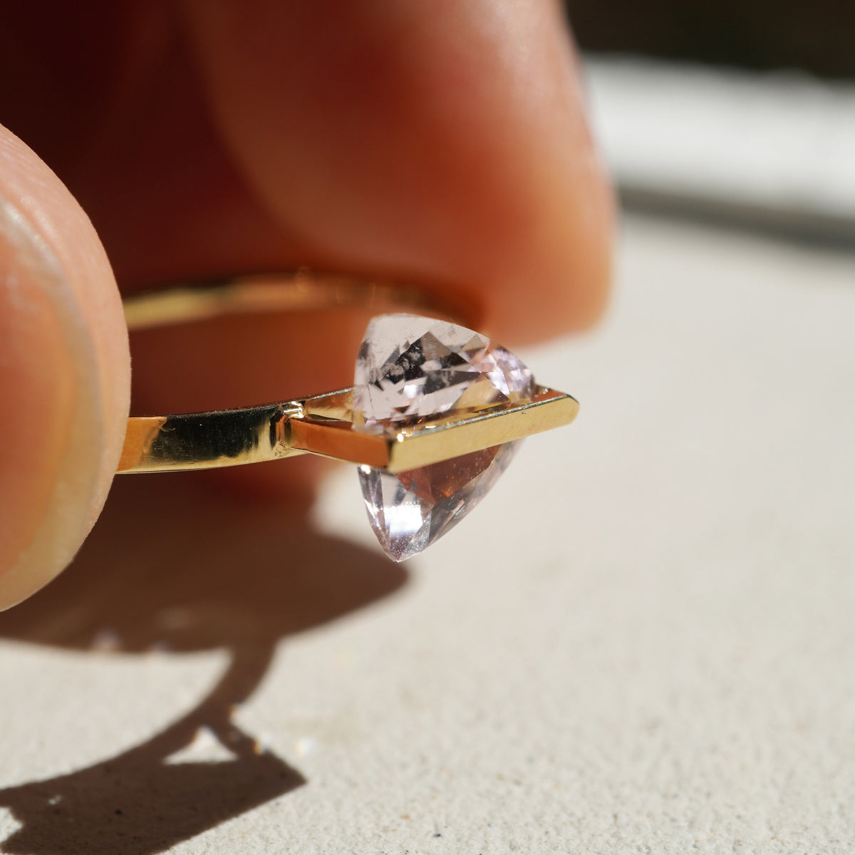 三角形の淡いピンク色のモルガナイトを10金を使ってセッティングしたバンドシリーズの指輪を指で摘んで自然光を石の中に通してみた様子