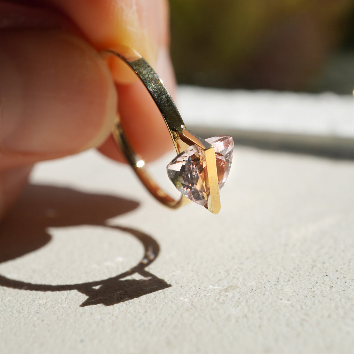 三角形の淡いピンク色のモルガナイトを10金を使ってセッティングしたバンドシリーズの指輪を指で摘んで自然光を石の中に通してみた様子。輝いているモルガナイトと、その影。