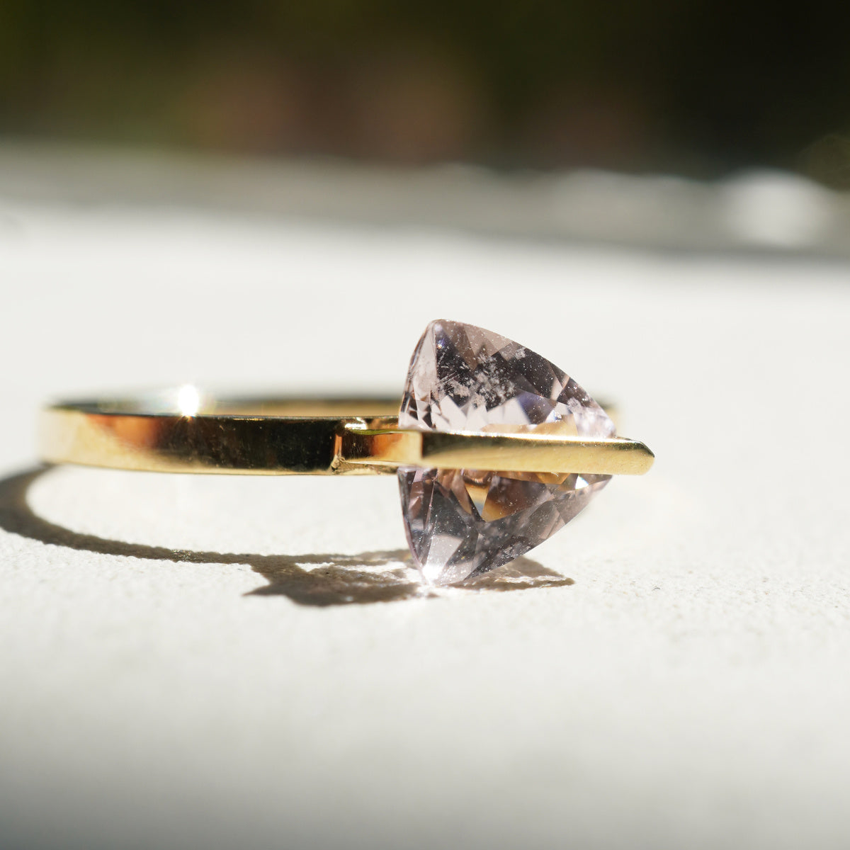 三角形の淡いピンク色のモルガナイトを10金を使ってセッティングしたバンドシリーズの指輪を自然光の中で斜めからアップで見た様子